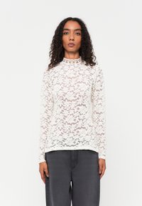 Fehér csipkéből készült, hosszú ujjas felső, magas nyakkal. Aprólékos virágminták díszítik. Sötétszürke nadrággal párosítva. Texturált és áttetsző dizájn.