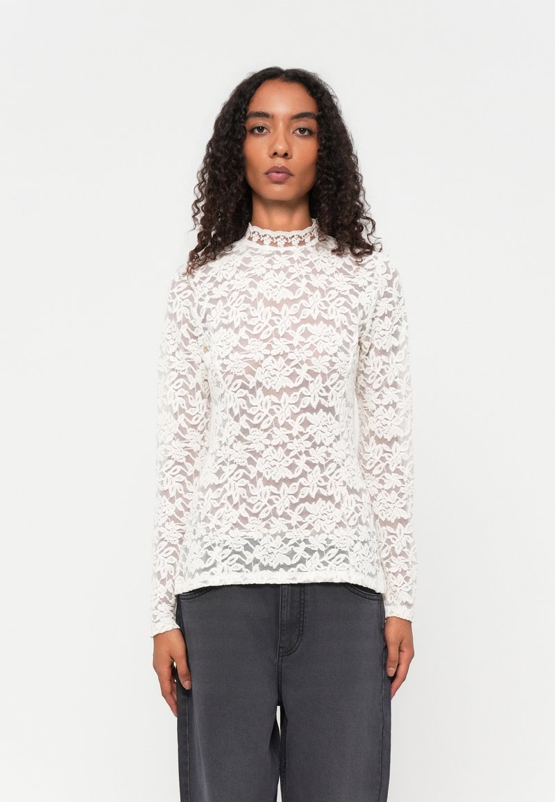 Fehér csipkéből készült, hosszú ujjas felső, magas nyakkal. Aprólékos virágminták díszítik. Sötétszürke nadrággal párosítva. Texturált és áttetsző dizájn.