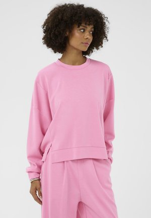 Göndör hajú nő laza illeszkedésű rózsaszín pulóverben és hozzáillő nadrágban, egyszínű világos háttér előtt áll.