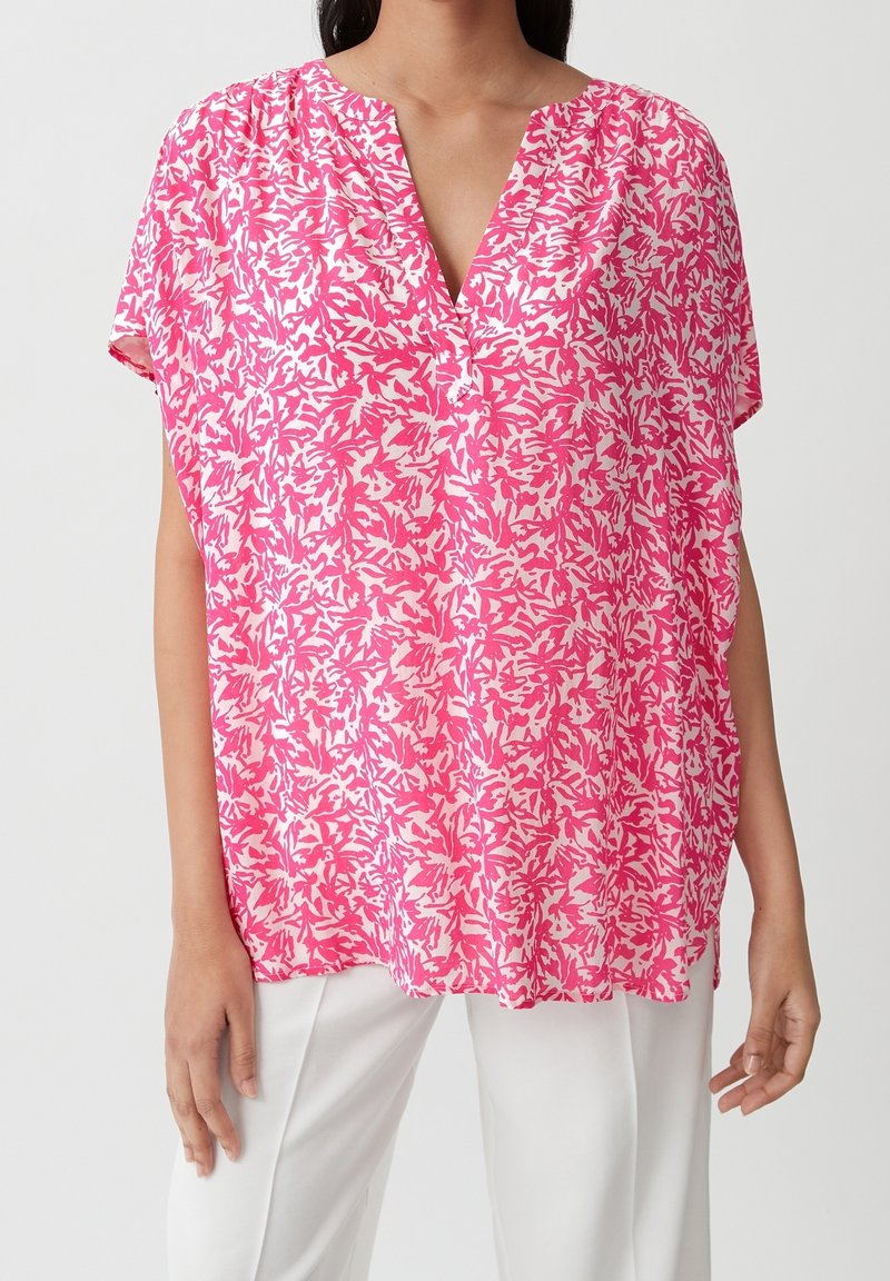 Femme portant un chemisier ample à manches courtes avec motif floral rose et blanc, col en V, et un pantalon blanc, sur fond uni.