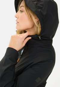 Sort hættetrøje med lynlås, struktureret stof, justerbar hætte og logoakcent på ærmet. Sideprofil med hånd ved kraven.