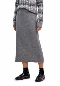 Person in einem grauen gestrickten Midirock, gemustertem grauen Pullover, schwarzen Socken und glänzenden schwarzen Slippern, stehend vor weißem Hintergrund.