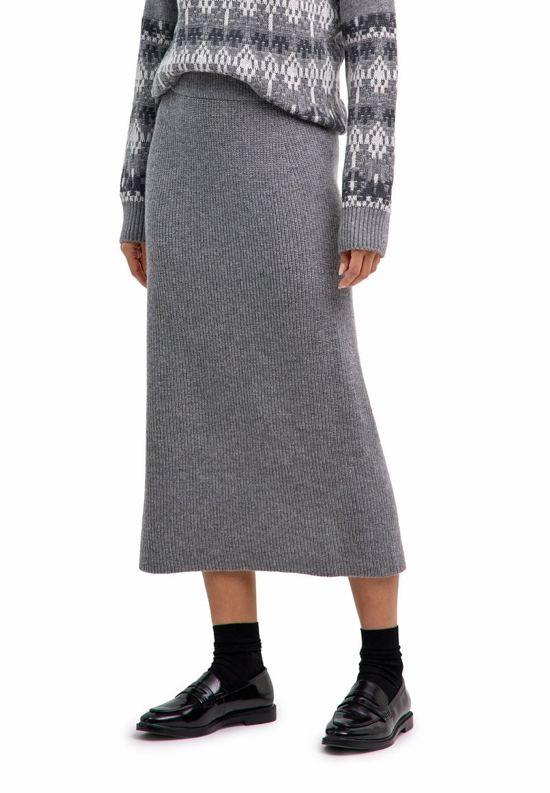 Person in einem grauen gestrickten Midirock, gemustertem grauen Pullover, schwarzen Socken und glänzenden schwarzen Slippern, stehend vor weißem Hintergrund.