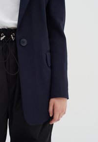 Námořnický blazer vyrobený z hladké tkaniny se zapínáním na jeden knoflík, přiléhavý střih a detail na přední kapse. Spárován s uvolněnými černými kalhotami.