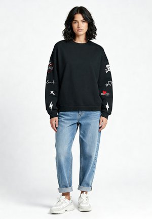 Felpa nera con dettagli ricamati sulle maniche, tra cui un cuore, un teschio e un fulmine. Abbinata a jeans azzurri arrotolati e sneakers bianche.