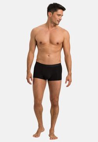 Black boxer shorts made of smooth fabric, featuring a grey elastic waistband. Model stands barefoot with a neutral pose against a white background.