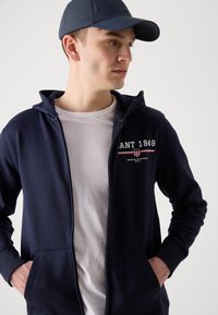 Sweat à zip bleu marine avec un logo sur le devant, des poches kangourou et une capuche à cordon, porté sur un t-shirt blanc. Casquette bleue avec un motif texturé.