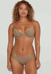 Sieviete ar cirtainiem matiem, tērpta brūnos svītrainos peldkostīmā, stāv pretī vienkāršai gaiši pelēkai fonā.