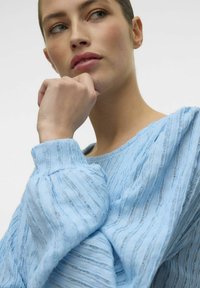 Heled sinise tekstuuriga pluus, millel on vertikaalsed triibud, ümmargune kaelus ja puffse varrukad, mida kannab modell neutraalses taustas.