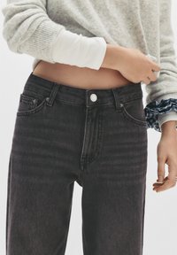 Personne portant un jean noir taille haute et un haut à manches longues en couches, exposant le ventre, avec une main ajustant l'ourlet du pull.