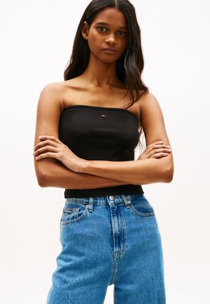 Ung kvinne med langt mørkt hår står med armene i kors, iført en svart stroppeløs topp og blå høyhalsede jeans mot en ensfarget bakgrunn.