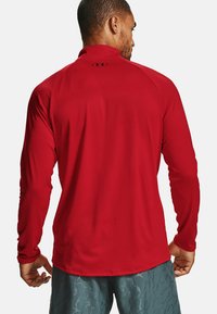 Rød langærmet træningsskjorte med høj krave og sort logo på ryggen. Glat tekstur, tætsiddende design, båret med mønstrede shorts.