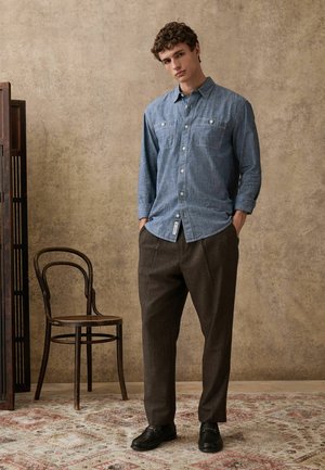 Denim-Hemd in Hellblau mit zwei Brusttaschen, kombiniert mit braunen, maßgeschneiderten Hosen und schwarzen Schuhen, vor einem neutralen Hintergrund.