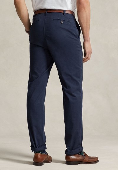 Man draagt marineblauwe broek, bruine leren riem, gebreide sokken en bruine leren loafers, staand met de rug naar de camera op een neutrale achtergrond.