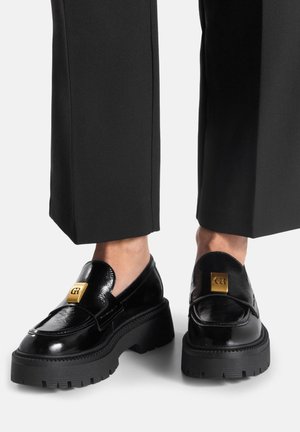 Schwarze Loafers aus Lackleder mit einer klobigen Plateausohle. Sie verfügen über einen goldfarbenen Akzent am Vorderfuß und eine glatte Textur.