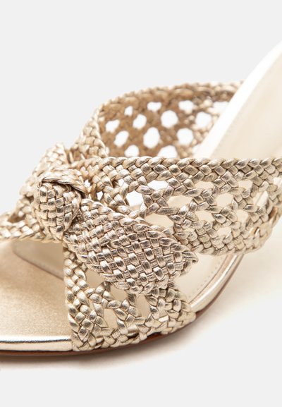 Μεταλλική χρυσή υφασμάτινη λουρίδα σανδαλιού με λεπτομέρεια δεμένης φιόγκος πάνω από ανοιχτό δάχτυλο σε φωτεινό φόντο.