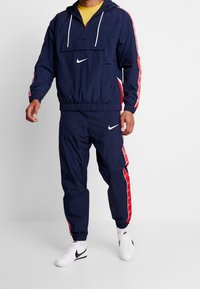 Marinblå Nike träningsdräkt med en halvmatchning zip-tröja med vit logotyp och röda sidostråkar, tillsammans med matchande byxor med elastisk midja.