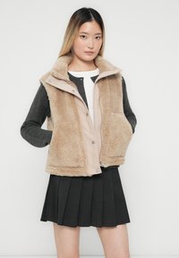 Vrouw in een beige donzige vest over een donkergrijze cardigan en een witte top, gecombineerd met een zwarte geplooide rok, staand tegen een witte achtergrond.