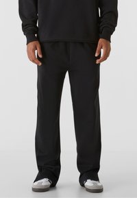 Pantaloni negri de trening cu croială lejeră, realizați dintr-un material moale. Prezintă o talie elastică și buzunare laterale. Purtați cu adidași albi și gri.