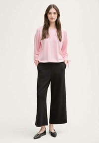 Blusentop in Hellrosa mit langen Ärmeln und gerippter Textur, kombiniert mit hochgeschnittenen, weiter geschnittenen schwarzen Hosen. Schwarze flache Schuhe runden den Look ab.