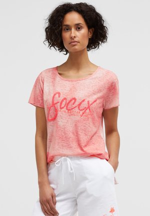 Woman with curly hair wearing a light pink short-sleeve shirt with text and white drawstring pants against a plain background.