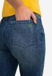 Jeans de mezclilla en azul oscuro con rayas verticales, con un bolsillo trasero y un pequeño detalle bordado en verde. La tela parece ligeramente texturizada.