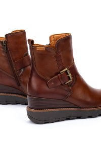 Bottes chelsea en cuir marron avec un talon compensé, présentant des détails tressés alternés et une fermeture éclair sur le côté. Sangle avec une boucle en métal en accent.