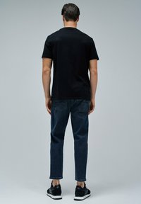 Hombre de pie de espaldas, vistiendo una camiseta negra lisa, jeans de mezclilla oscura y zapatillas negras y blancas, sobre un fondo gris.