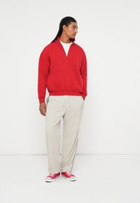 Pulover roșu cu fermoar pe jumătate, pantaloni bej cu dungi laterale și adidași roșii. Material neted, croi lejer. Modelul este orientat spre stânga, pe un fundal neutru.