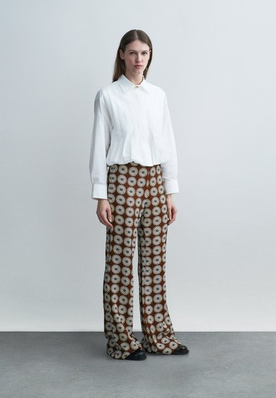 Νεαρή γυναίκα που φοράει λευκό χαλαρό πουκάμισο με κουμπιά και καφέ παντελόνι με φαρδιά μπατζάκια και λευκά λουλουδάτα σχέδια, στέκεται σε γκρι πάτωμα μπροστά σε ανοιχτόχρωμο τοίχο.