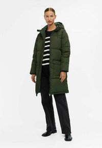 Groene gewatteerde jas met een capuchon, met een rits aan de voorkant, zijzakken en een gewatteerde textuur. Gedragen over een zwart-wit gestreept topje en zwarte broek.