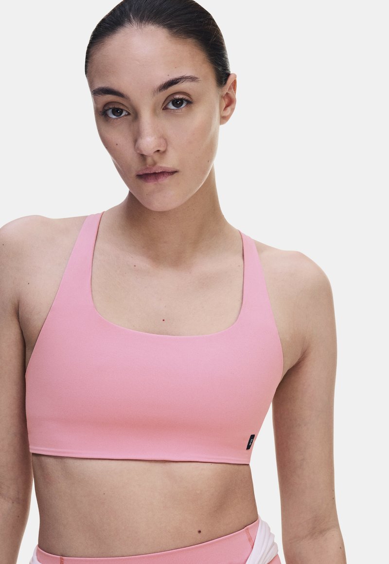 Femme aux cheveux foncés tirés en arrière, portant un soutien-gorge de sport rose clair sur un fond blanc uni.