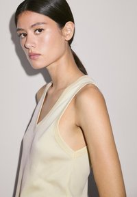 Ärmelloses Top mit V-Ausschnitt aus leicht gelbem Rippstoff. Verfügt über breite Schulterträger und eine taillierte Silhouette. Glatte Textur, minimalistisches Design.