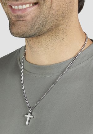 Silberne Kettenhalskette mit einem metallischen Kreuzanhänger, getragen über einem grauen T-Shirt. Nahaufnahme der Halskette und des Halsbereichs.