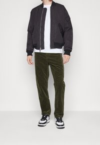 Svart bomberjacka, vit T-shirt, gröna manchesterbyxor och svart-vita sneakers. Jackan har dragkedja och ribbade ärmslut.