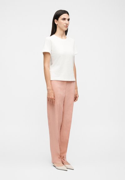 Γυναίκα όρθια, φοράει λευκό πουκάμισο με κοντά μανίκια, ροζ παντελόνι με ψηλή μέση και μπεζ παπούτσια με μυτερή μύτη σε απλό φόντο.