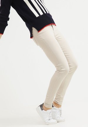 Osoba se naklání dozadu, má na sobě béžové úzké kalhoty, bílé tenisky s leopardím potiskem a tmavě modrý svetr s bílými pruhy a červeným lemováním.