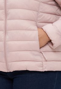 Svetloružová puffová bunda s prešívanou textúrou, so zipsom vpredu a bočnými vreckami. Tkanina sa zdá byť hladká a ľahká.