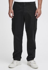 Schwarze Cargo-Hosen aus weichem, strapazierfähigem Stoff, mit Seitentaschen, einer lockeren Passform und subtilen Nähdetails. Kombiniert mit weißen Sneakers.