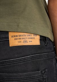 Jeans oscuros de mezclilla con una etiqueta de cuero que dice "DENIM DRIVEN SINCE 1989" y "CRAFTING QUALITY GARMENTS." Camisa verde olivo por encima.