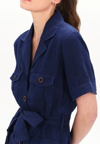 Vestido de lino azul marino con mangas cortas, dos bolsillos en el pecho y una cintura ceñida con cinturón. Presenta detalles de botones y un cuello con muesca.