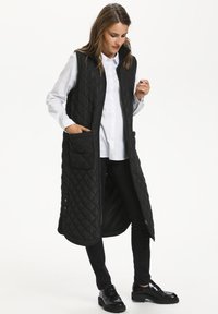 Gilet long noir matelassé avec une fermeture éclair sur le devant, un col montant, des poches latérales et une texture lisse, porté sur une chemise blanche et un pantalon noir.