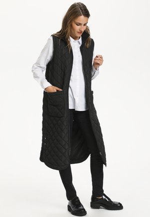Gilet long noir matelassé avec une fermeture éclair sur le devant, un col montant, des poches latérales et une texture lisse, porté sur une chemise blanche et un pantalon noir.