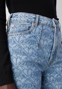 Light blue denim jeans with a geometric pattern, high-waisted design, contrasting stitching, and a small pink accent on the button.