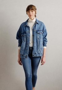 Jeansjakke i lys blå med to brystlommer, knappeåpninger og oversized passform, kombinert med tettsittende blå jeans og hvit genser med høy hals.