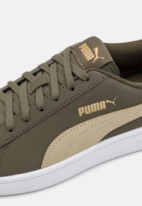 Sneaker verde oliva con accenti beige, sovrapposizioni testurizzate e logo Puma dorato. Presenta chiusura con lacci e suola in gomma bianca.