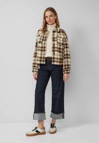 Geruite jas in crème, bruin en zwart, gecombineerd met een crèmekleurige coltrui en donkere jeans. Sneakers hebben zwarte en crèmekleurige accenten met gumzolen.