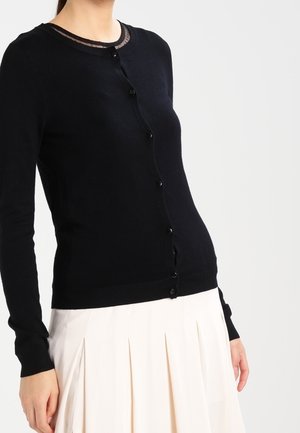 Frau trägt einen taillierten schwarzen, geknöpften Cardigan mit transparenten Details am Ausschnitt und einen plissierten weißen Rock.