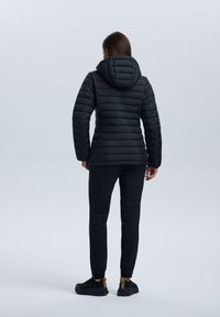 Frau, die von hinten steht, trägt eine schwarze gesteppte Kapuzenjacke, schwarze Hosen und schwarze Sneakers mit sandfarbenen Akzenten vor einem einfarbigen hellen Hintergrund.