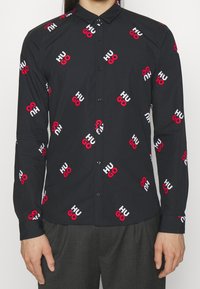 Czarna koszula na guziki z powtarzającym się nadrukiem logo "HUGO" w kolorach czerwonym i białym, wykonana z gładkiej tkaniny, z klasycznym kołnierzykiem i długimi rękawami.